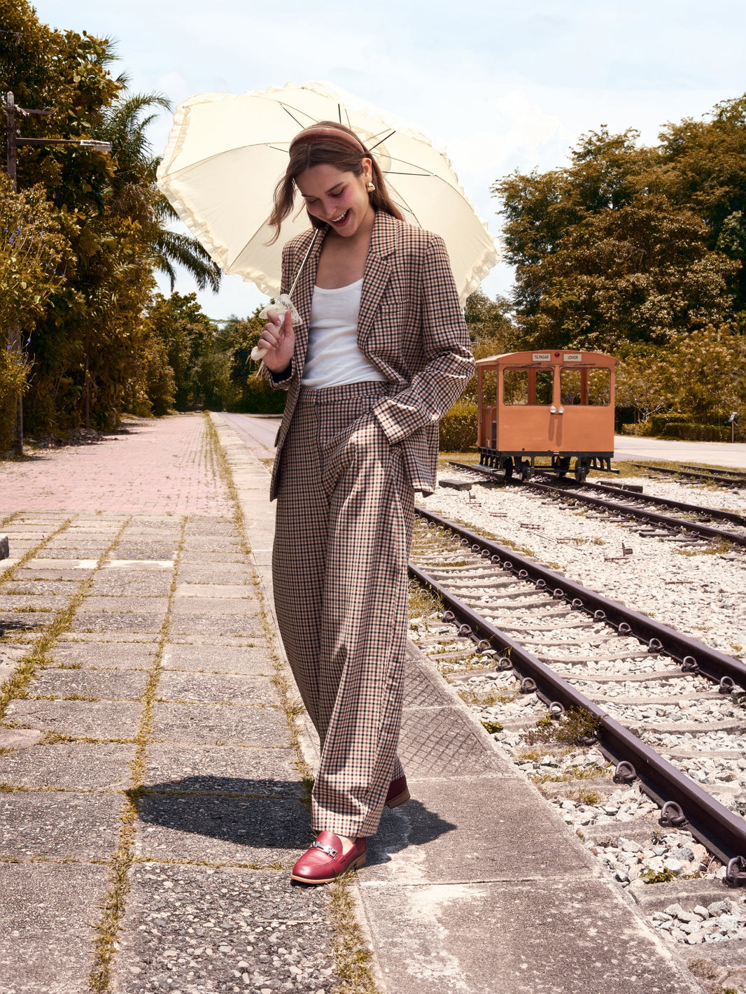 Woman in a checked suit holding a white umbrella walks by railway tracks, wearing prettyFIT Nova Loafers in burgundy.