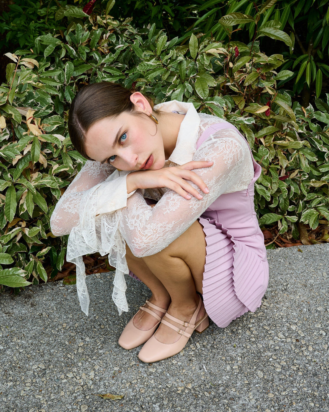 Model in a lace blouse and pleated pink dress wearing prettyFIT Mary Jane heels in nude, seated outdoors with lush greenery in the background.